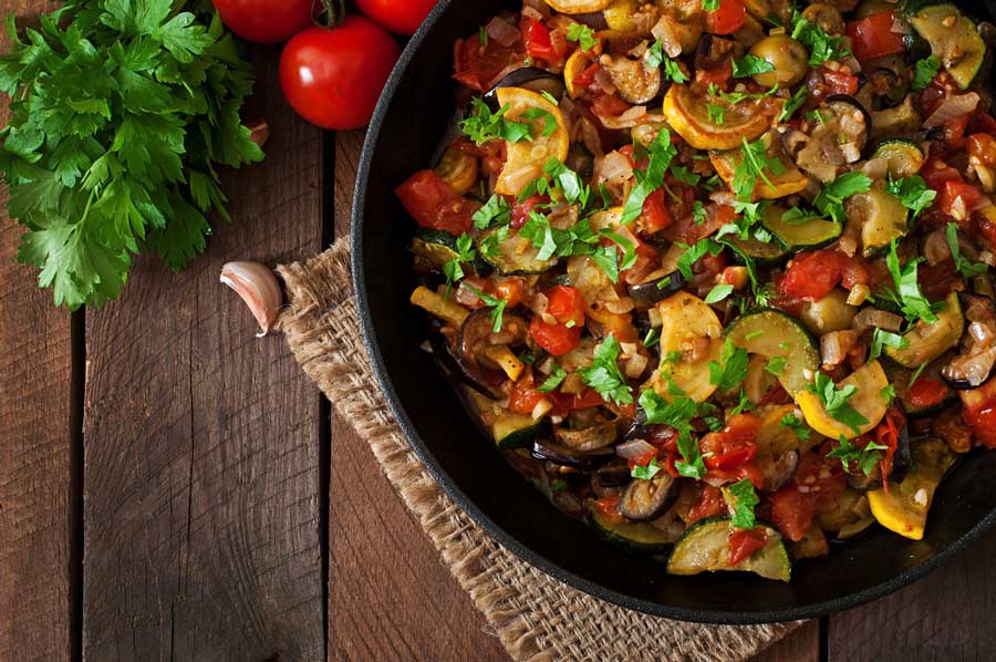 Готовий рататуй у вигляді овочевого рагу, посипаний зеленню, у глибокій сковороді на дерев’яному фоні з петрушкою та томатами.