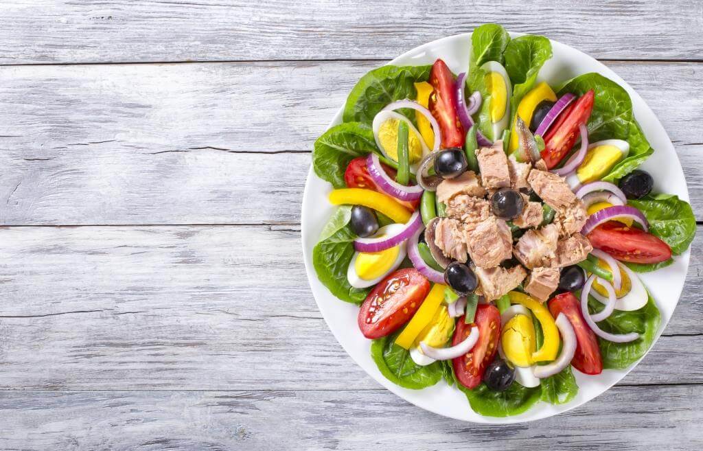 Салат Нісуаз з тунцем, яйцем, оливками, овочами та зеленню на білому дерев’яному фоні