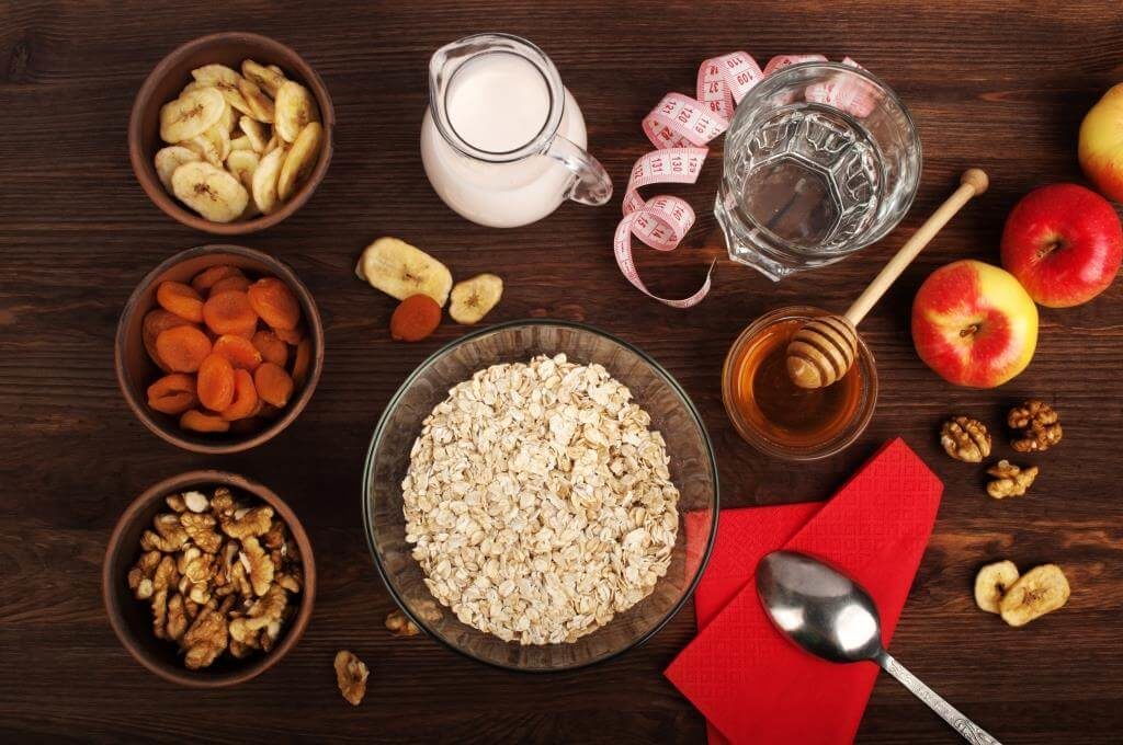 Стильна композиція для сніданку: вівсянка в мисці, оточена сушеними бананами, курагою, волоськими горіхами, медом, яблуками і молоком.