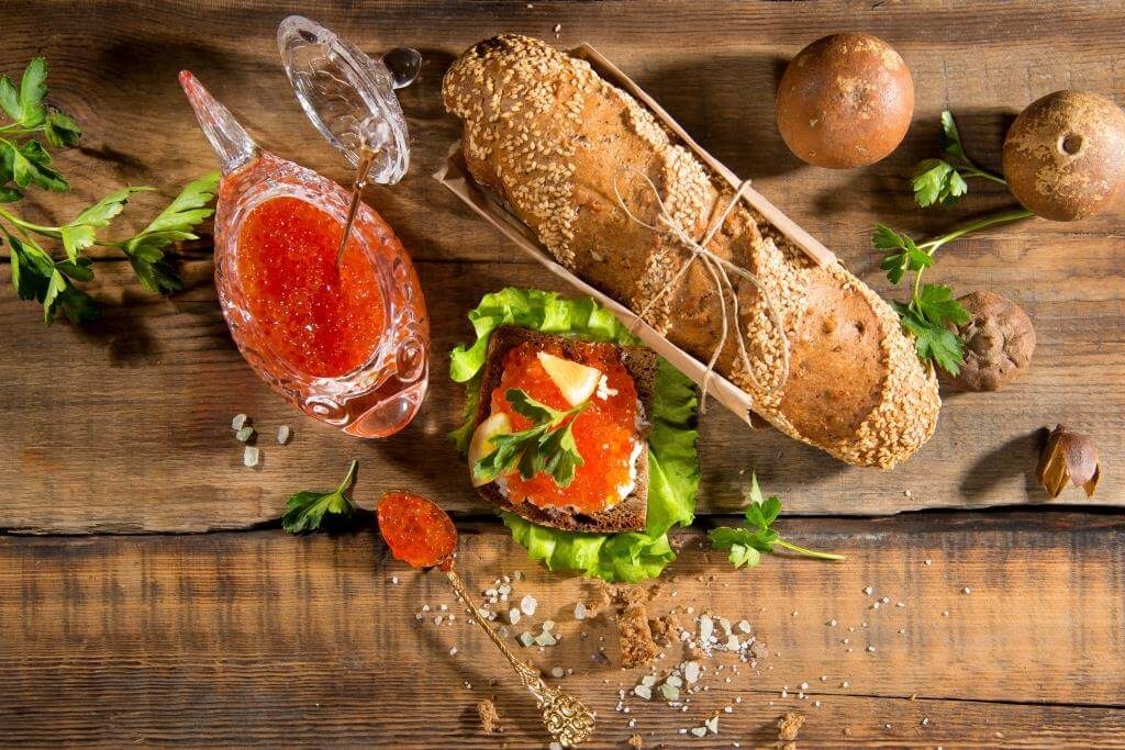 Хліб з насінням, червона ікра в соусниці та сервірування з зеленню на дерев'яному столі.