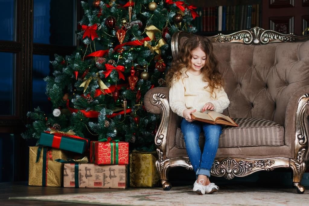 Дівчинка читає книжку поруч з ялинкою і новорічними подарунками.