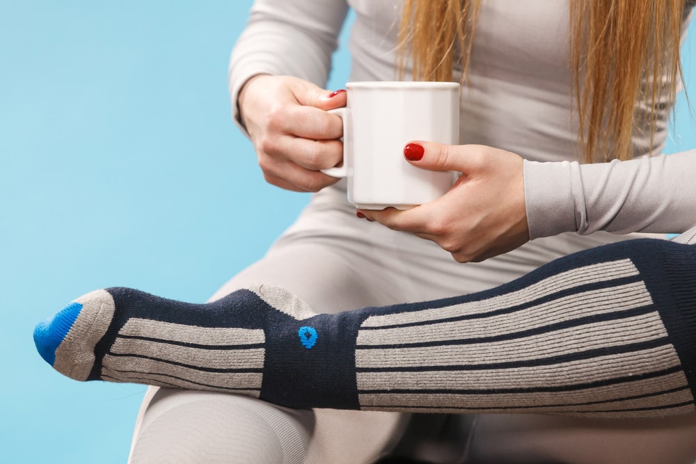 Жінка у термобілизні тримає горнятко, сидячи у теплих зимових шкарпетках