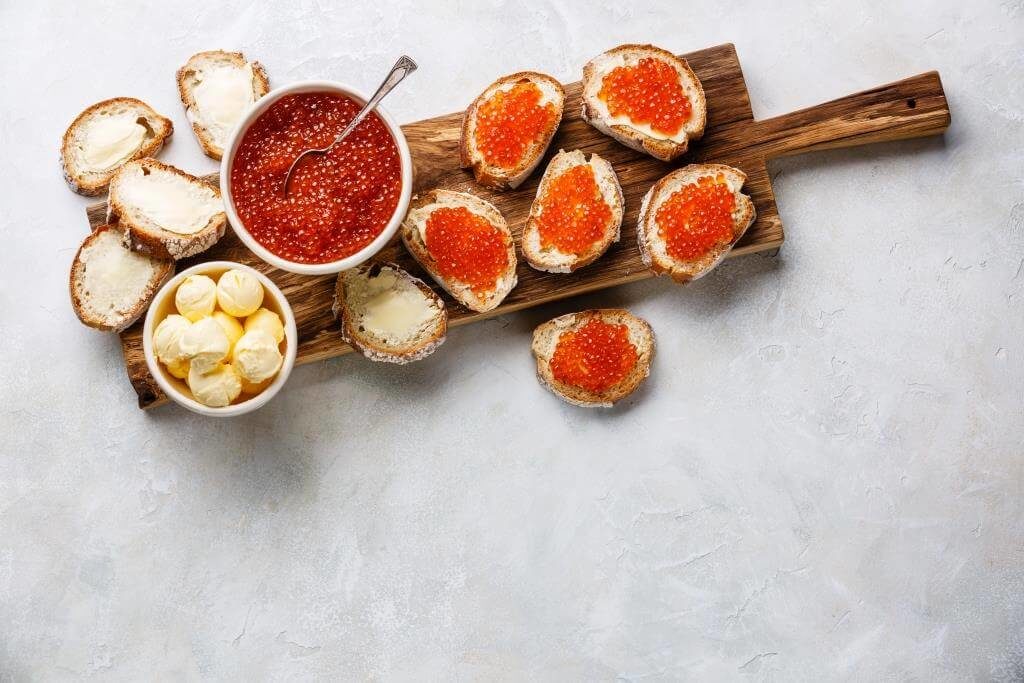 Нарізаний хліб із маслом, червона ікра в мисці та готові бутерброди на дерев’яній дошці.