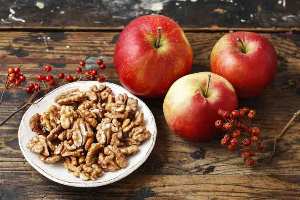 Стиглі червоні яблука та тарілка з волоськими горіхами на дерев’яному фоні.