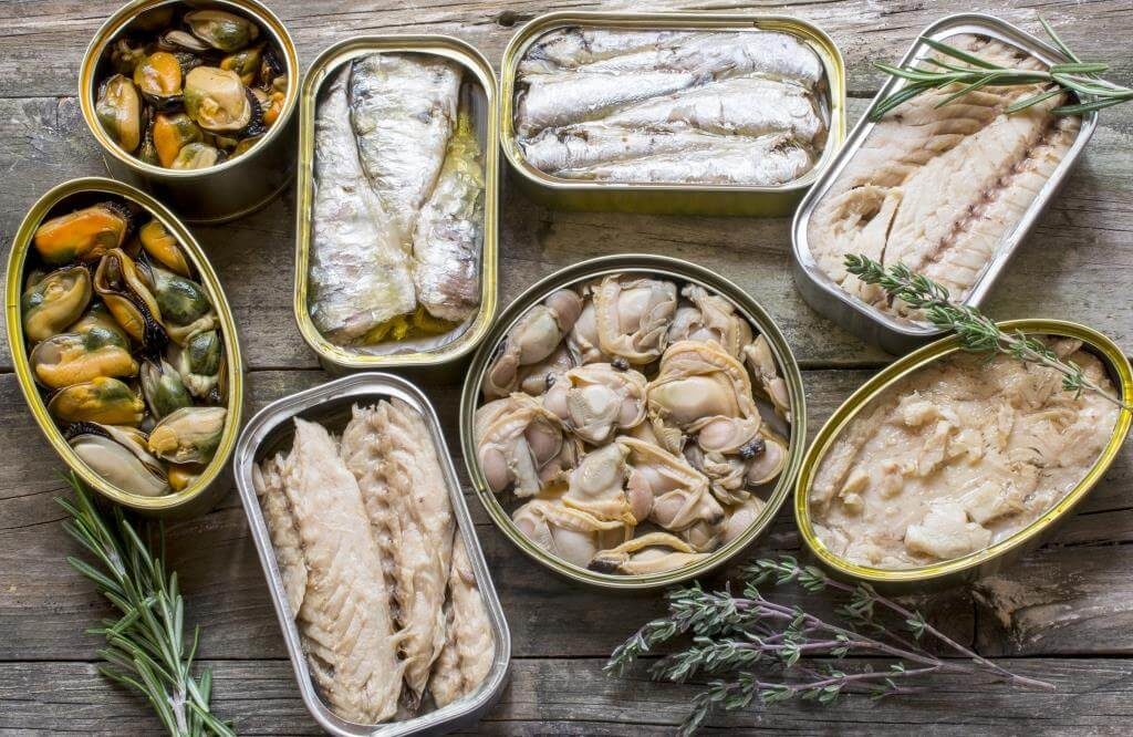 Різні види рибних і морепродуктових консервів у відкритих банках на дерев’яному столі.
