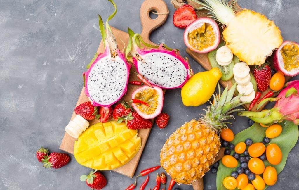 Асорті з екзотичних фруктів: пітахая, маракуя, манго, ананас, полуниця, лимон, банани, ківі, чорниця і кумкват на сірому фоні