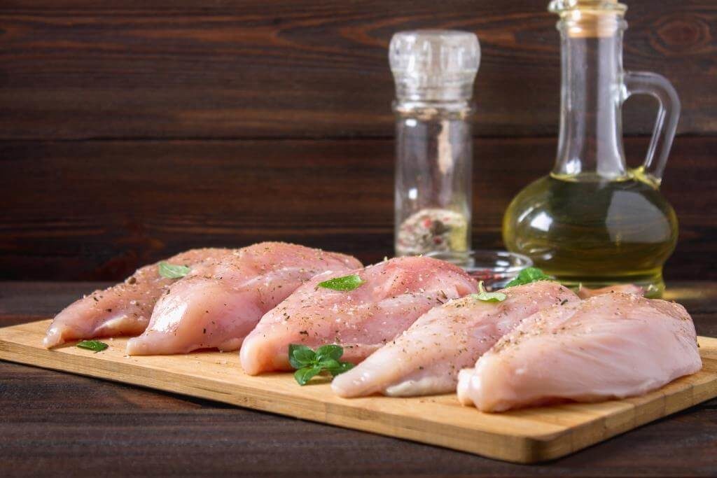 Сирі курячі філе на дерев’яній дошці, приправлені спеціями та травами, поруч пляшка олії та млинок для перцю