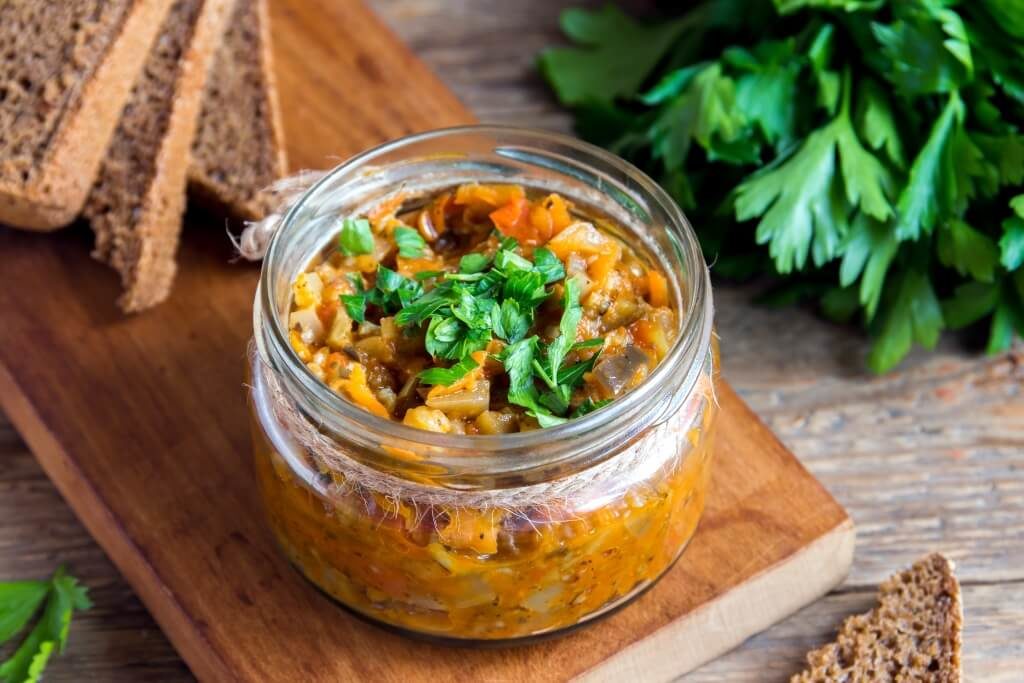 Овочева ікра в банці, прикрашена зеленню, поруч із житнім хлібом