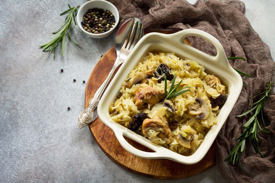 Печена капуста з грибами, чорносливом і м’ясом у керамічній формі на дерев’яній підставці, з розмарином і перцем.
