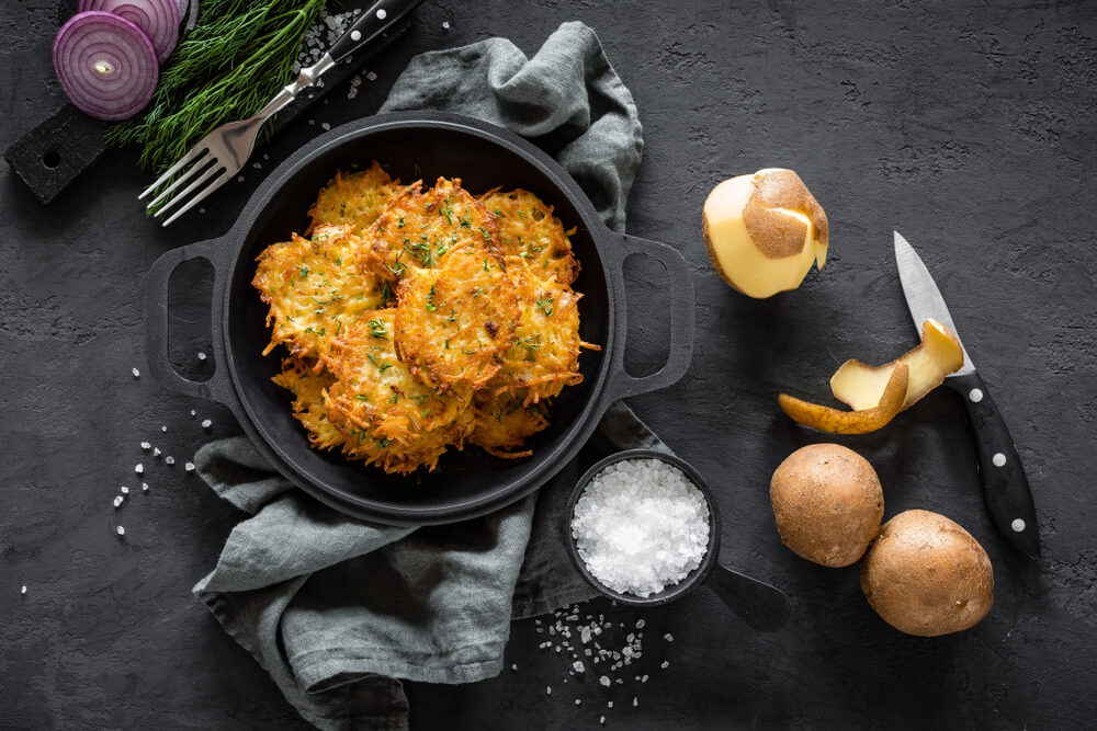 Драники з картоплі у чорній сковороді, поруч сирі картоплини, ніж і сіль.