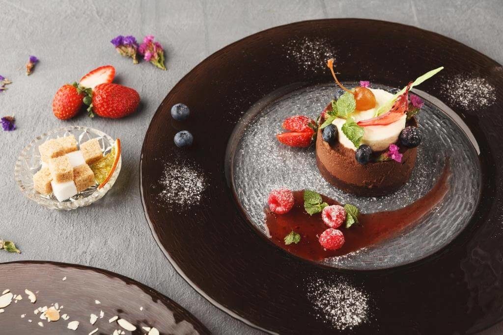 Десерт на шоколадній основі з кремовим топінгом, ягодами і листям м’яти, на чорній скляній тарілці з цукровою пудрою.