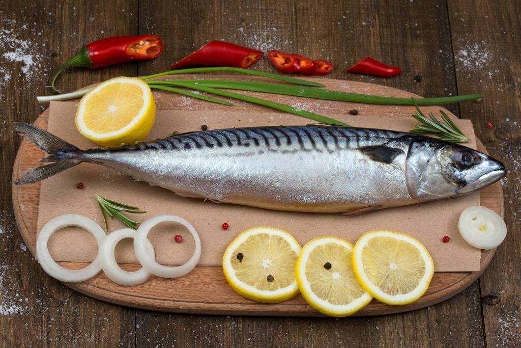 Скумбрія на дерев’яній дошці, оточена скибками лимона, кільцями цибулі, чилі, зеленню та перцем